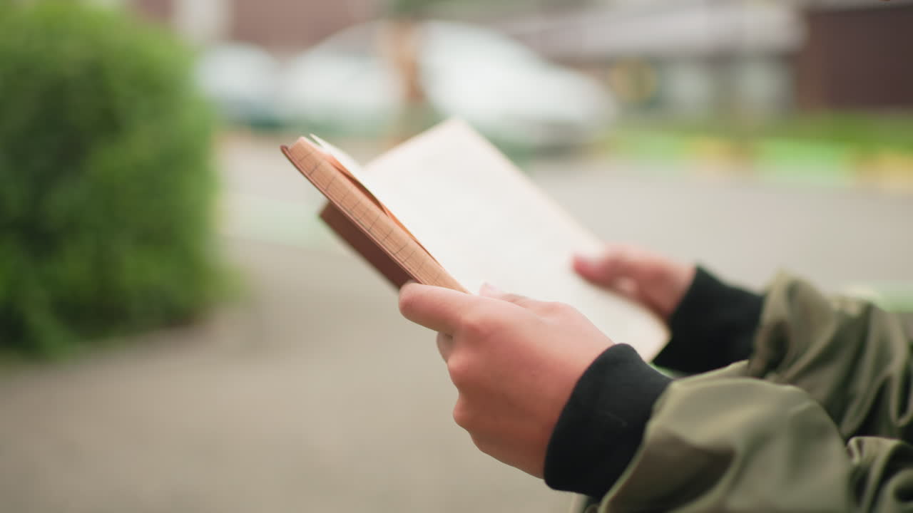Primo piano di una mano che tiene un libro e sfoglia le pagine all'aperto, con sfondo sfocato in cui si vedono un edificio e un'auto parcheggiata