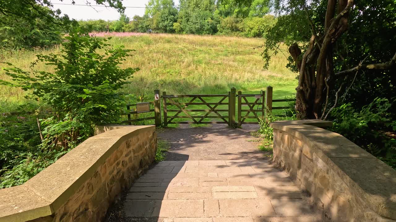 Camera moves steadily down a sunlit stone path, crossing a small bridge and approaching a wooden gate in a lush botanical garden setting