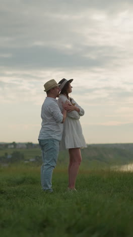un momento tierno entre dos amantes, como un hombre con un sombrero, camisa blanca y vaqueros sostiene a una mujer con un vestido blanco en una colina cubierta de hierba al atardecer. la escena irradia calor, amor y conexión