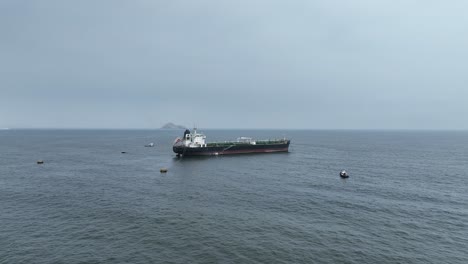 aerial forward dolly shot of a large cargo ship with smaller boats floating in the pacific ocean with view of hills from an island in the background