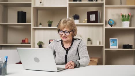 Una-Mujer-Mayor-Con-Gafas-Inicia-Una-Videollamada-Con-Una-Sonrisa-Usando-Su-Computadora-Portátil-Y-Envía-Un-Beso-Al-Aire