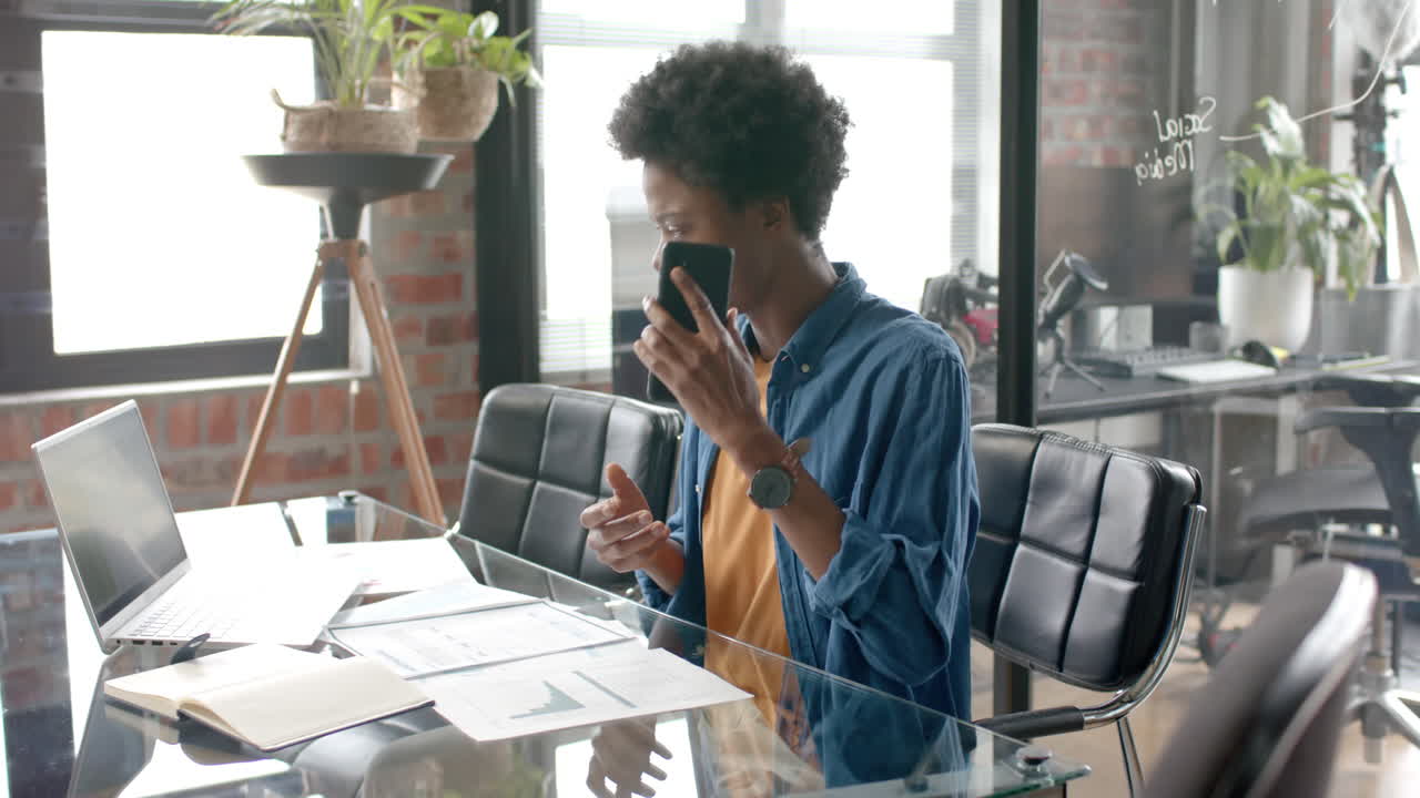 Afroamerikansk afslappet forretningsmand sidder ved skrivebordet og taler i smartphone derhjemme, i slow motion.