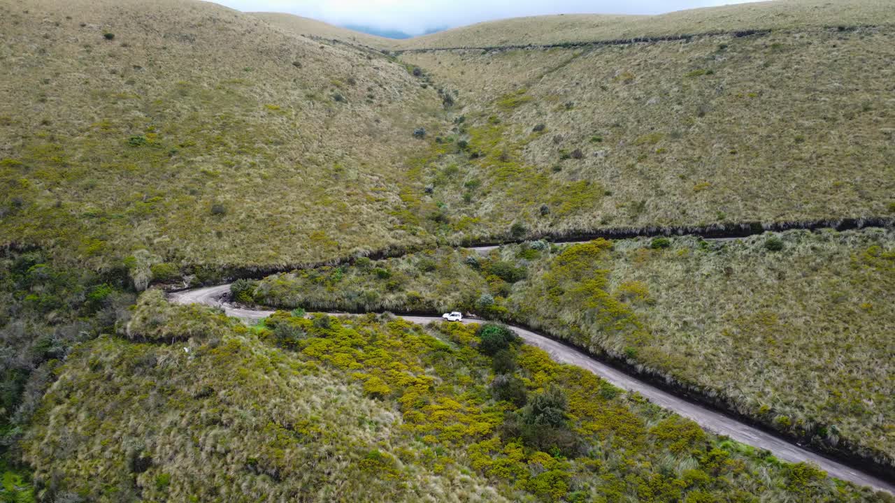 Aerial dolly-out shot capturing a lone car navigating a winding dirt road in the vast Ecuadorian paramo. As the drone pulls back, the vehicle fades into the immense highland landscape.