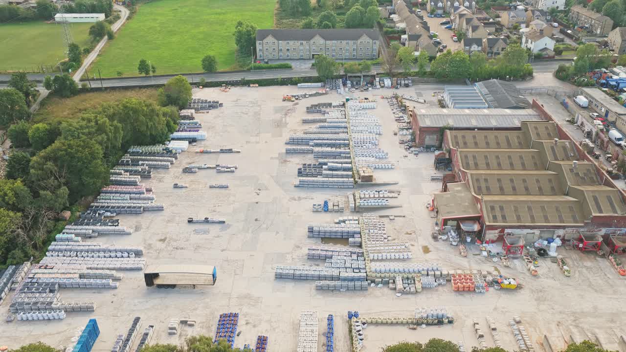 Drone pulls out to show Sunderland Way industrial park’s large material storage yard, warehouses, and adjacent housing set beside green space in Brighouse, West Yorkshire