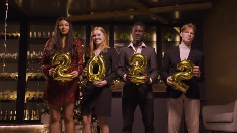 group of friends wearing elegant clothes holding balloons with the numbers 2022 while dancing in new year's party