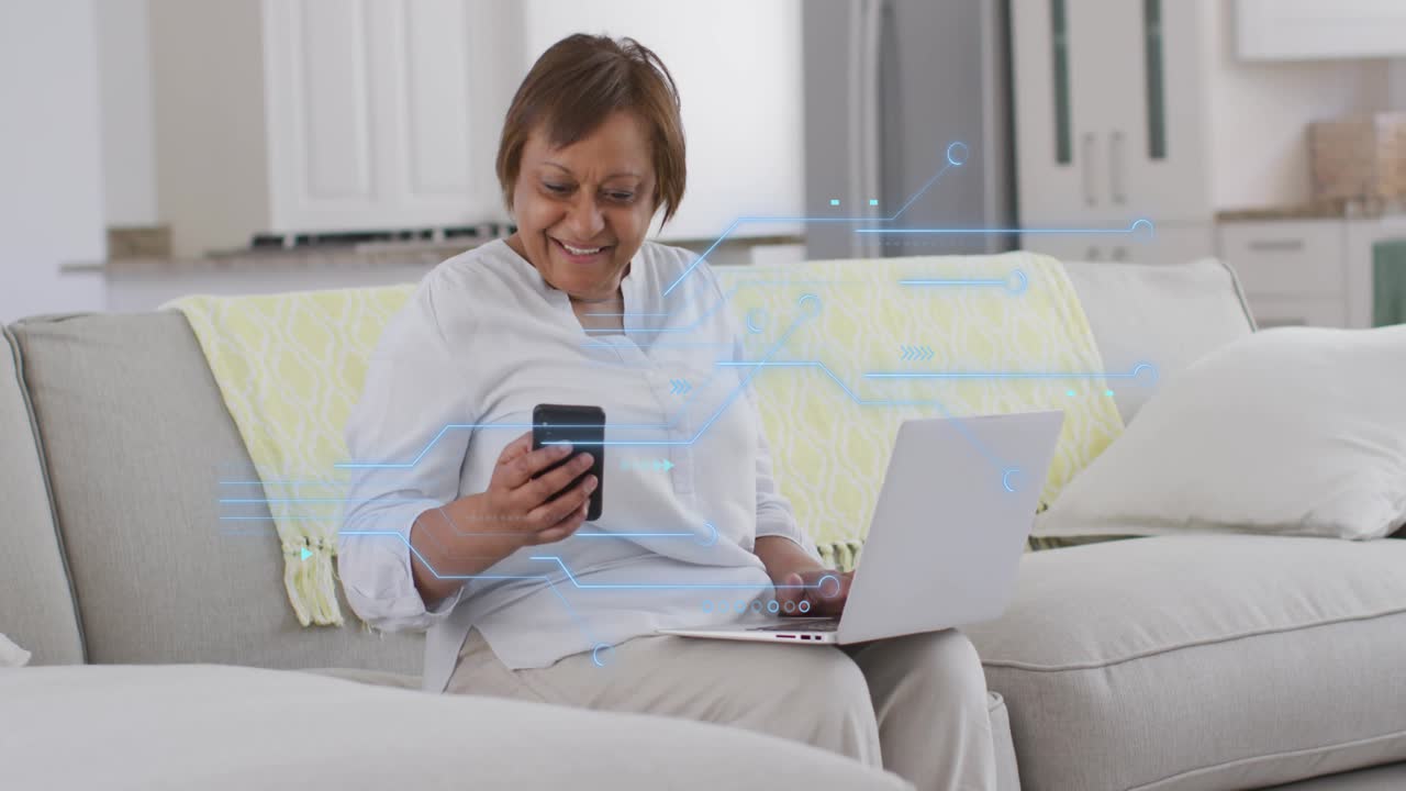 Senior woman sitting sofa typing laptop and tapping phone, causing HUD overlay animating tech data