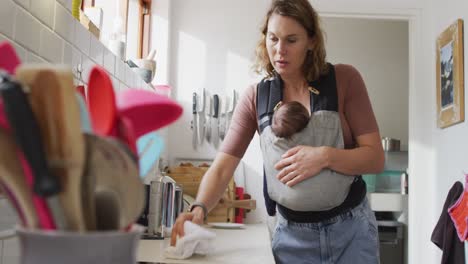 Video-of-caucasian-mother-with-newborn-baby-in-baby-carrier-cleaning-kitchen