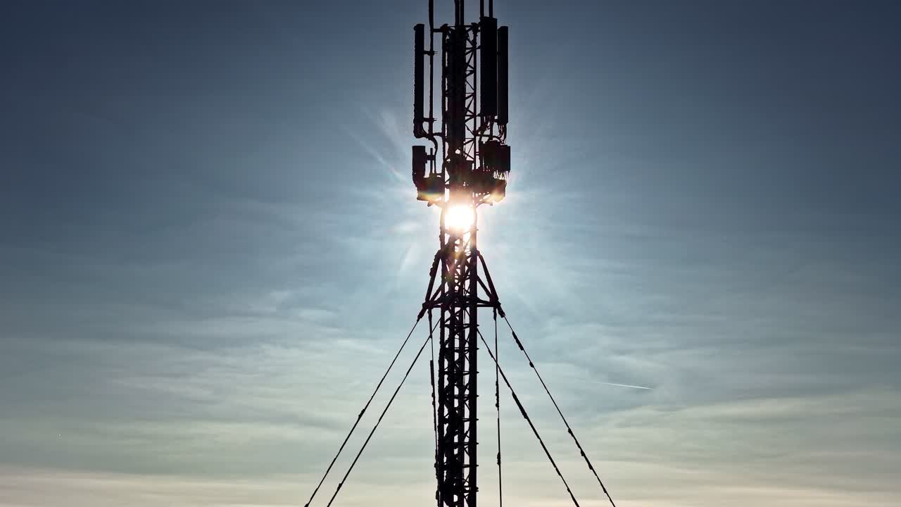 Drone che scende verticalmente accanto a un alto palo per telecomunicazioni, con il sole perfettamente allineato dietro le antenne che crea un bagliore intenso contro un cielo azzurro limpido.
