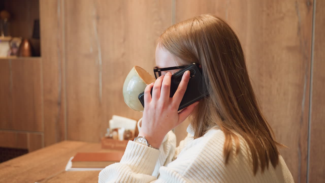 side view young woman wearing glasses sitting in cozy cafe holding phone near ear engaged in call while having breakfast with books and napkin holder on wooden table