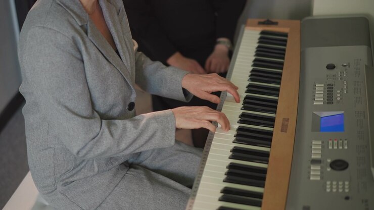 Donna che suona il pianoforte con l'insegnante, studentessa in abito grigio concentrata sui tasti, coach che guida il posizionamento delle mani, piccolo studio di scuola di musica, spartito sul leggio, prova tranquilla e pratica della tecnica.