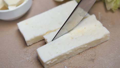 close-up of a knife cutting a block of white cheese on a cutting board