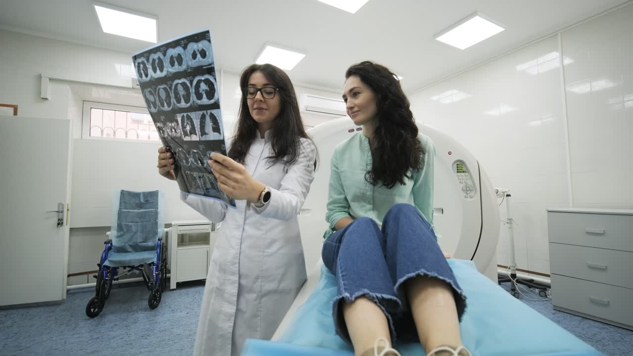 la radióloga explica los buenos resultados de la tomografía computarizada para la paciente joven, mostrando la instantánea con imágenes, observando y analizando la tomografia computarizada en una clínica moderna junto a una máquina de escáner computarizado moderna.