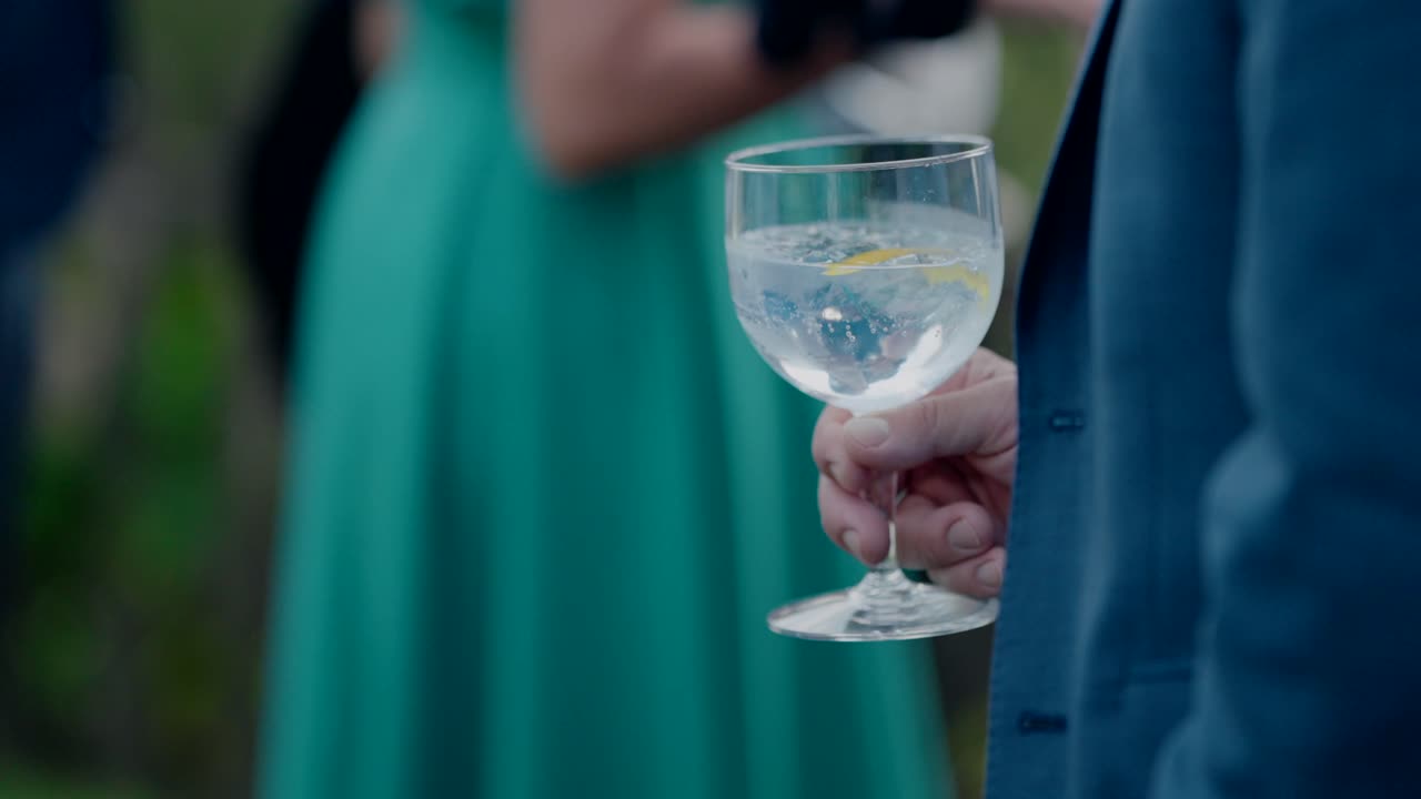 Close up of person holding clear cocktail with ice and lemon in blue jacket