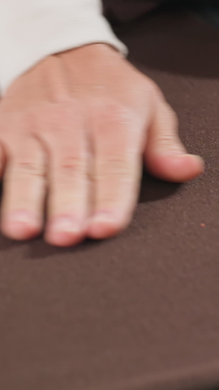 테이블 위에 놓인 갈색 천, 템플릿을 정렬하는 백인 손, 재단 준비를 위해 천을 접는 아틀리에 제작자, 클로즈업 주름 디테일, 핀 없는 정렬, 데님 배경, 의류를 위한 패턴 배치