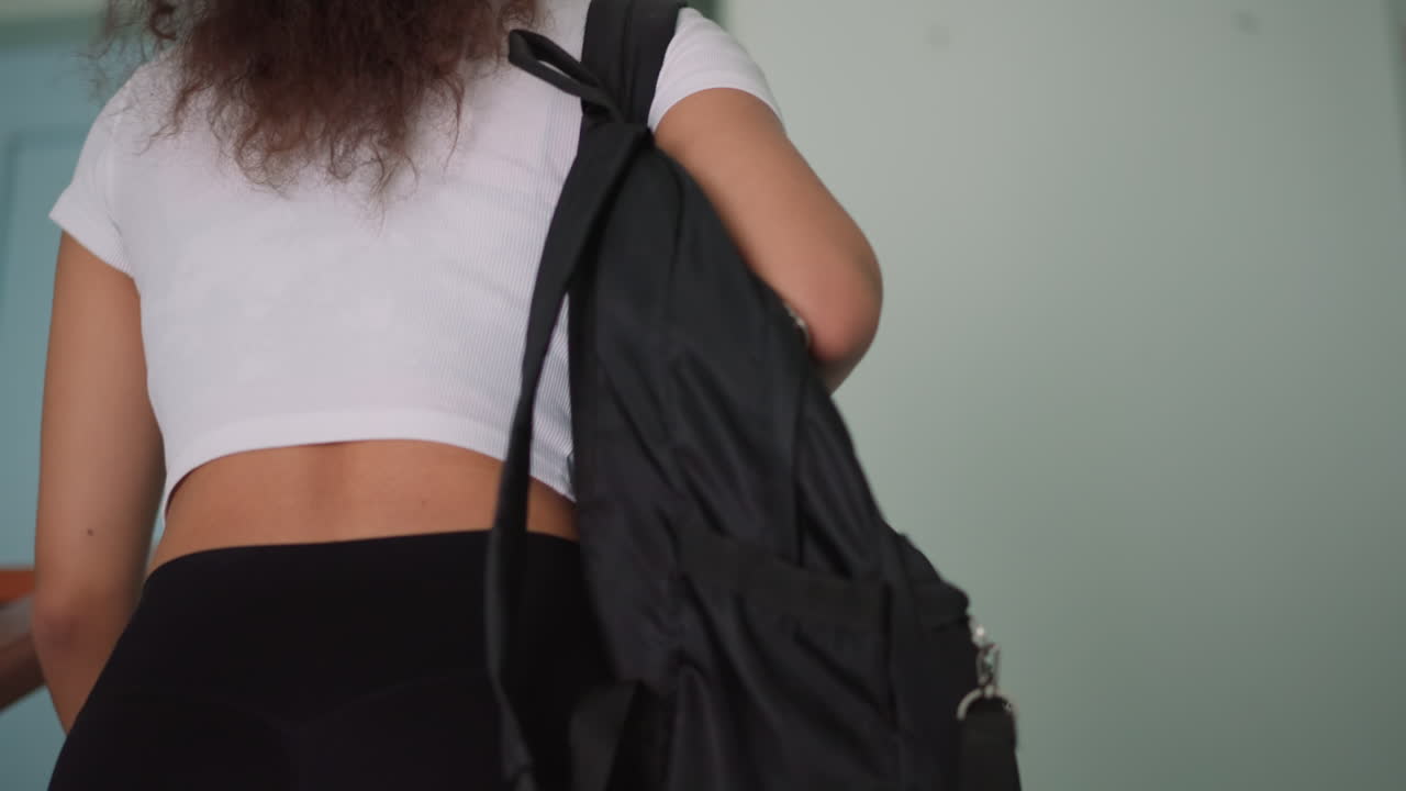 Back view of woman in casual white top and black leggings carrying school bag while walking calmly indoors, focus on relaxed movement and lifestyle detail, showing balance of study, fashion