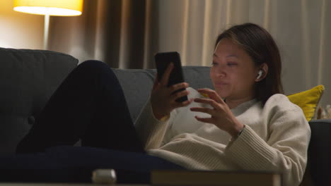 woman spending evening at home on sofa wearing wireless earbuds and streaming music or podcast from mobile phone 6