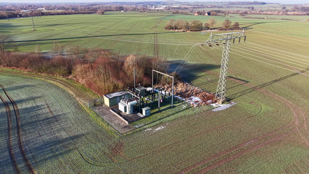 Vista aerea da drone di una sottostazione elettrica rurale collegata a un traliccio di trasmissione ad alta tensione, circondata da campi agricoli e vegetazione invernale. La scena evidenzia la rete elettrica fondamentale