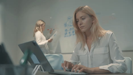 Joven-Mujer-De-Negocios-Escribiendo-En-Una-Computadora,-Colega-Trabajando-Con-Gráficos-Y-Diagramas-En-Una-Pizarra-Blanca