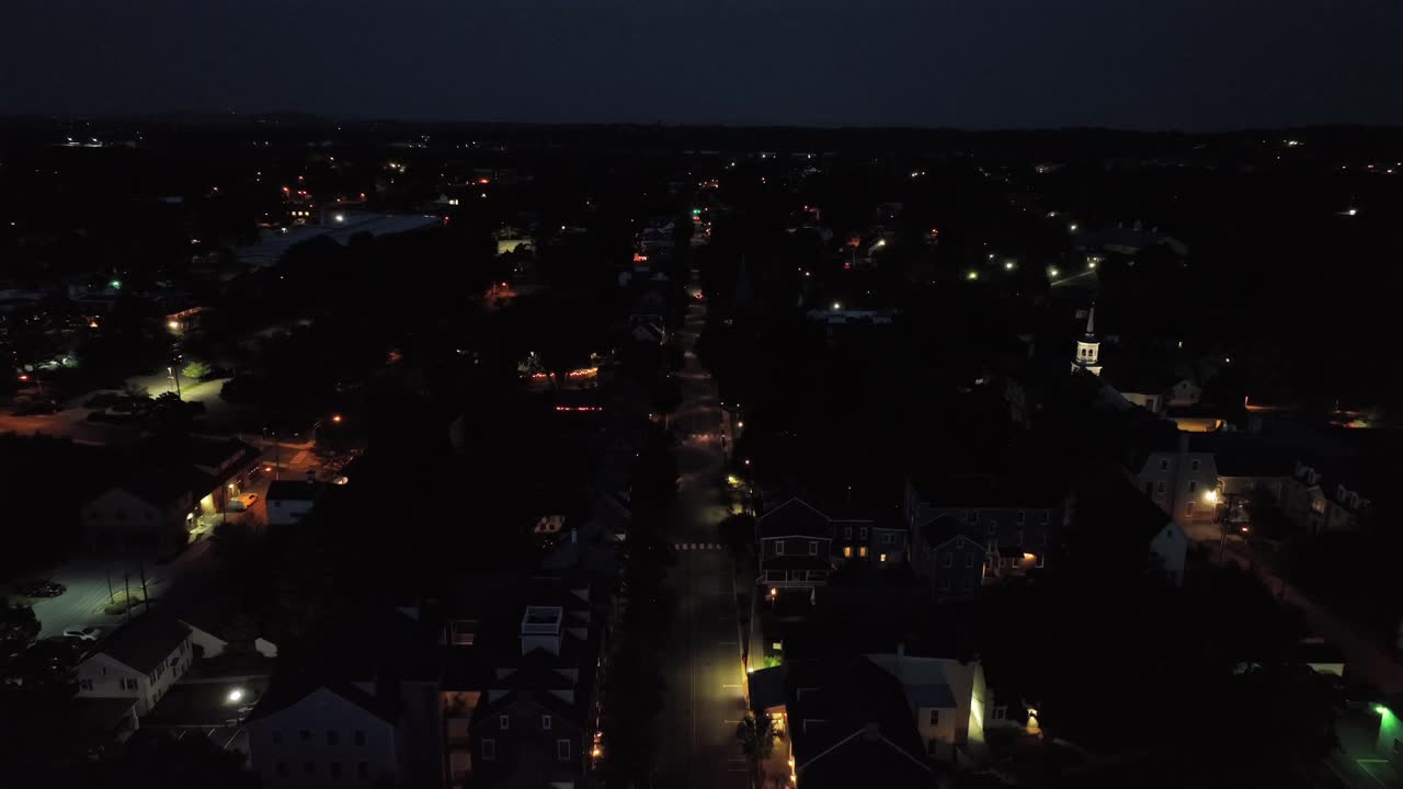Noční scéna v malém americkém městě s jedoucím autem na Hlavní ulici. Široký letecký záběr. Spící rodiny v domech. Osvětlené kostelní věže. Klidná obytná čtvrť.