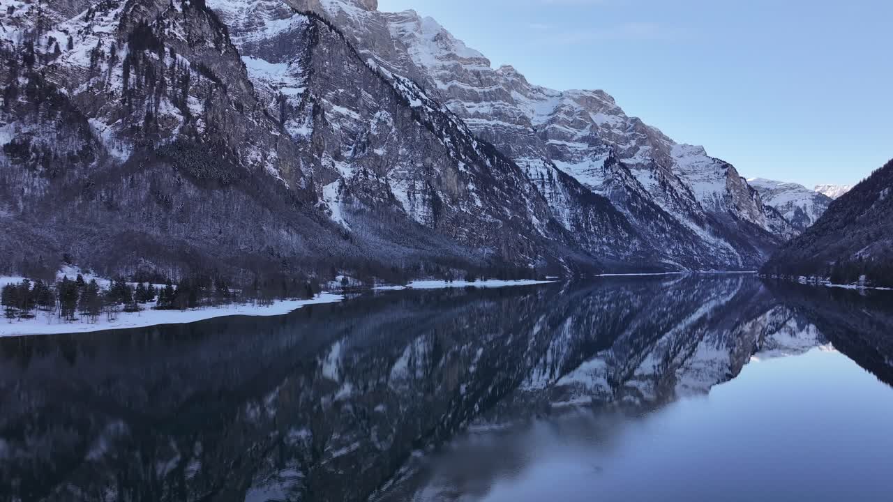 Aerial view of Klöntalersee, Glarus, Switzerland, showcasing a tranquil winter landscape with snow-capped mountains reflected on the calm lake waters. A serene and scenic alpine setting.