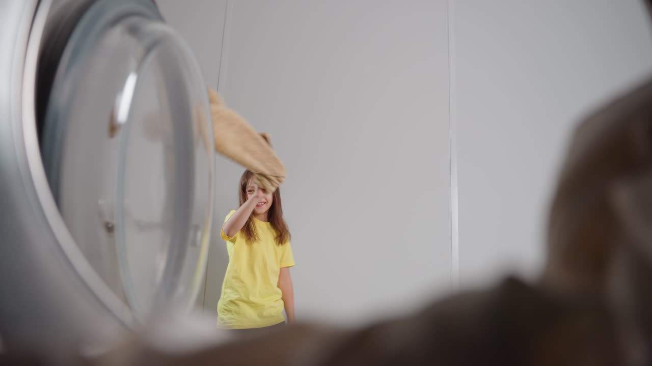 La vista interna di una lavatrice mostra una ragazza energica che fa roteare un panno, camminando sorridendo, porta aperta a sinistra, un leggero riflesso sul bordo del vetro, una lavanderia luminosa, movimento giocoso, tessuti puliti, faccende gioiose