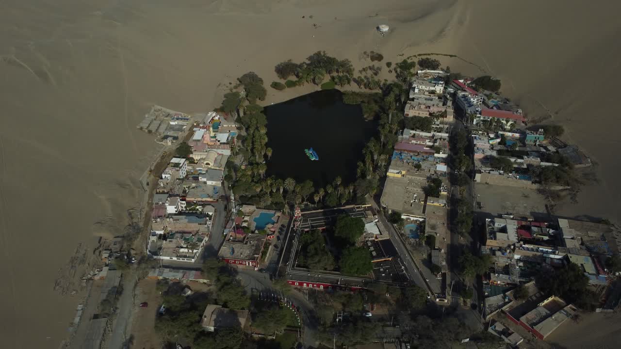 A small lagoon in the middle of the desert. Drone flies backwards tilting camera up revealing all the sand dunes in the horizon. Located in "Huacachina" in Ica, Peru.