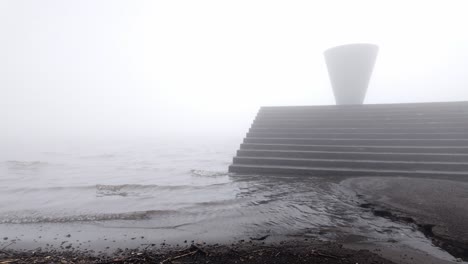 static wide shot of modern concrete staircase partly underwater at misty lakeshore, abstract cone sculpture above steps in thick fog and soft light