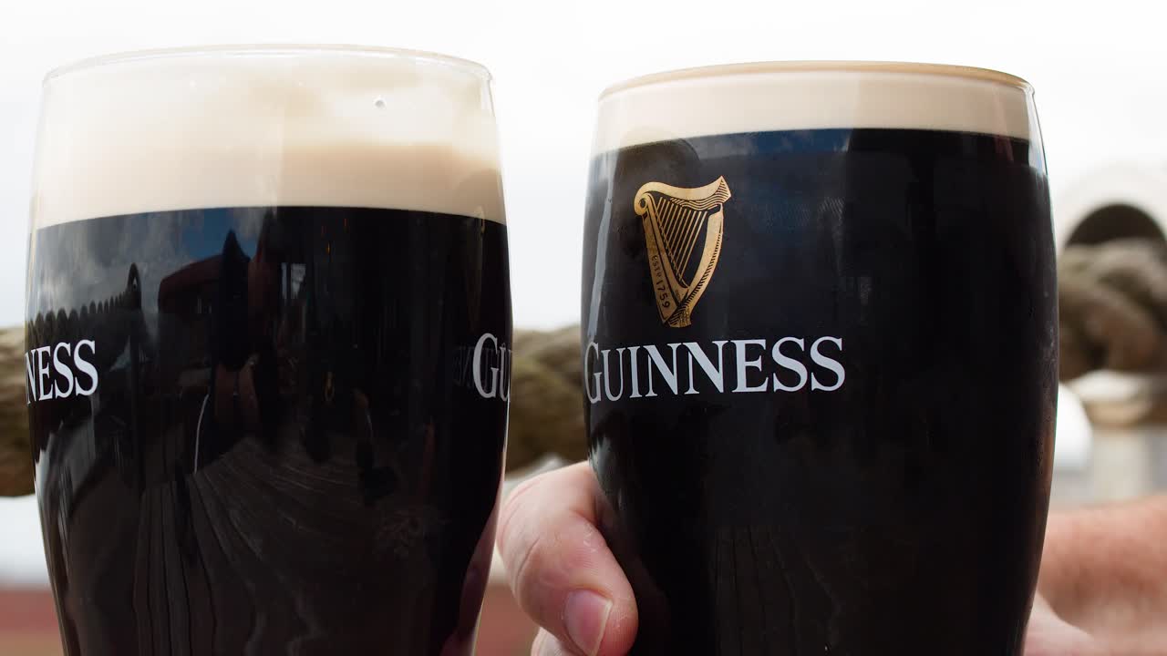 Two hands clink dark beer glasses outdoors, natural daylight, close-up, steady camera, casual mood