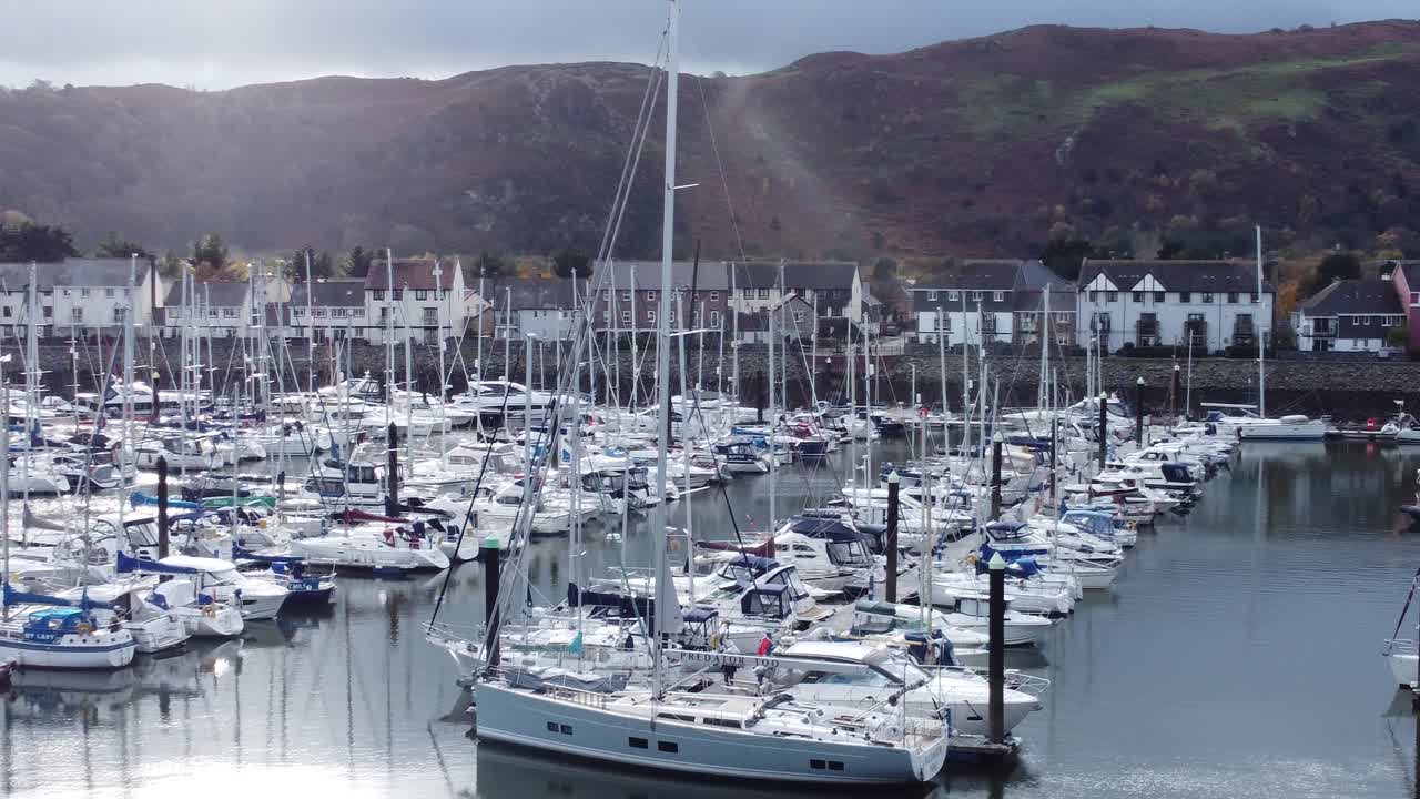Vista aerea panoramica e lussuosa di un villaggio di appartamenti sul lungomare del porto con yacht e barche a vela sotto la costa montuosa, ripresa in discesa verso destra.