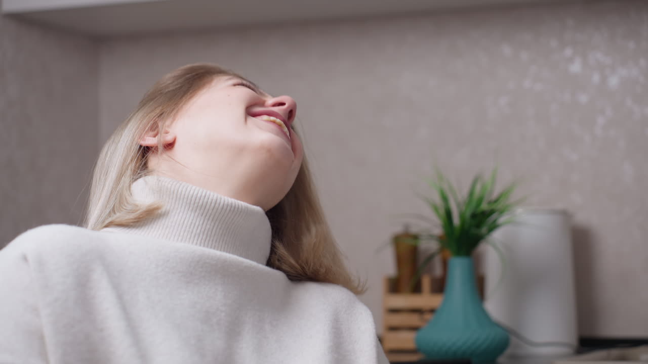 Smiling mother in white sweater laughing joyfully with warm expression during conversation at home kitchen table, showing happiness, affection, relaxed mood, family bonding moment, candid lifestyle