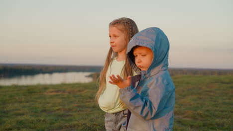 dos niños pequeños de pie en una colina con vistas a un río al atardecer.