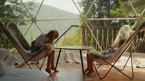 siblings sit in cozy woolen chairs in glamping camp overlooking forest under morning sun. family morning in apartment overlooking mountains. comfort and tranquility