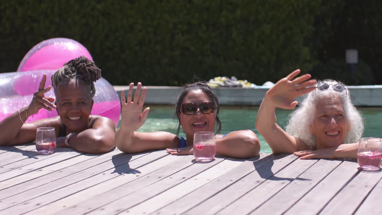 Mujeres diversas saludando tras ver la cámara, apoyadas en una terraza de madera con bebidas rosadas y gafas de sol.