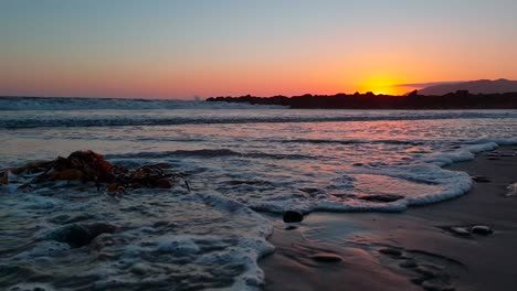 Tiro-De-Carro-Bajo-De-Algas-En-La-Playa-Durante-La-Puesta-De-Sol-Con-La-Luz-Del-Sol-Reflejándose-Bellamente-En-La-Arena-Y-Las-Olas-Que-Entran-En-La-Playa-Estatal-De-San-Buenaventura-En-Ventura,-California,-Ee.uu.