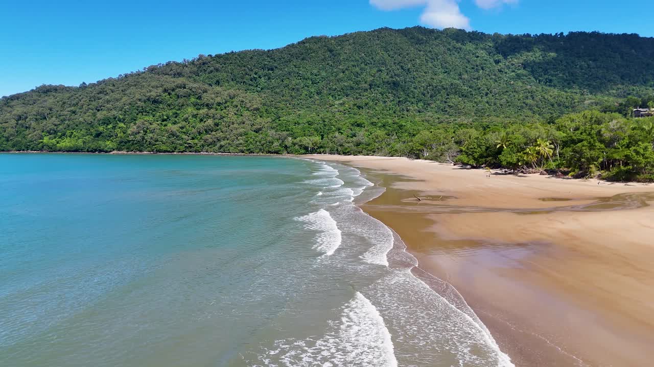 Drone footage glides above a deserted tropical beach with gentle waves, lush rainforest, and bright daylight, highlighting Cape Tribulation’s pristine coastline