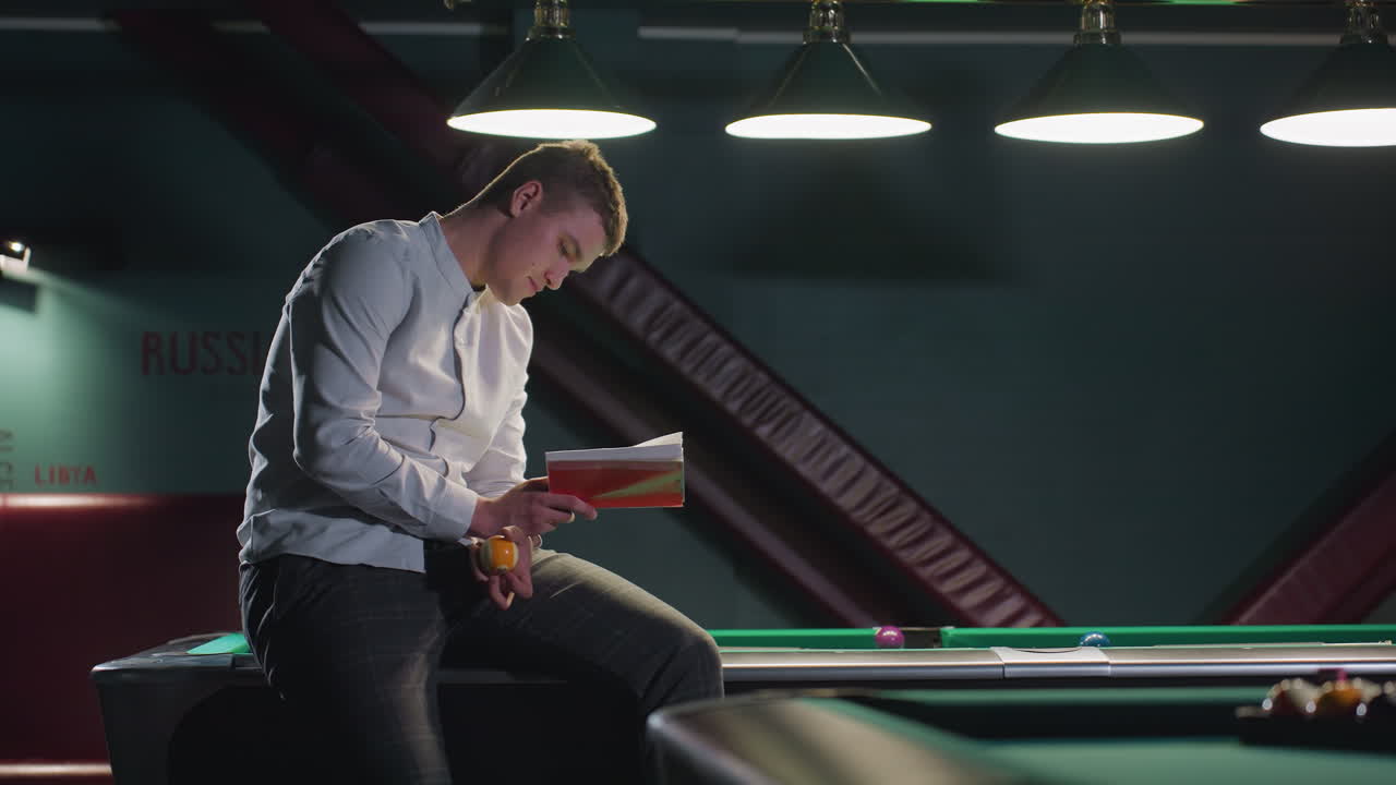 Una persona con un'espressione serena, indossa una camicia bianca e pantaloni a quadri, seduta su un tavolo da biliardo verde mentre legge un libro e fa rotolare una palla da biliardo in mano. Un altro tavolo da biliardo si trova sullo sfondo.