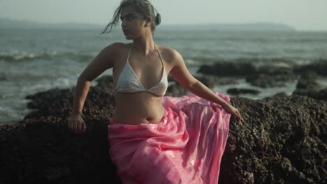 joven con camiseta blanca y falda rosa sentada en una roca junto al mar, viento en su cabello, estado de ánimo contemplativo, de día