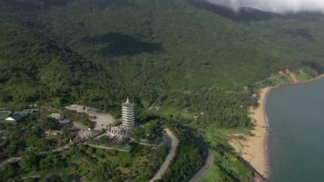 Aerial-view-tall-Lady-Buddha-statue-and-temple-tower-with-huge-mountains,-stunning-coastline-and-ocean-in-Da-Nang,-Vietnam