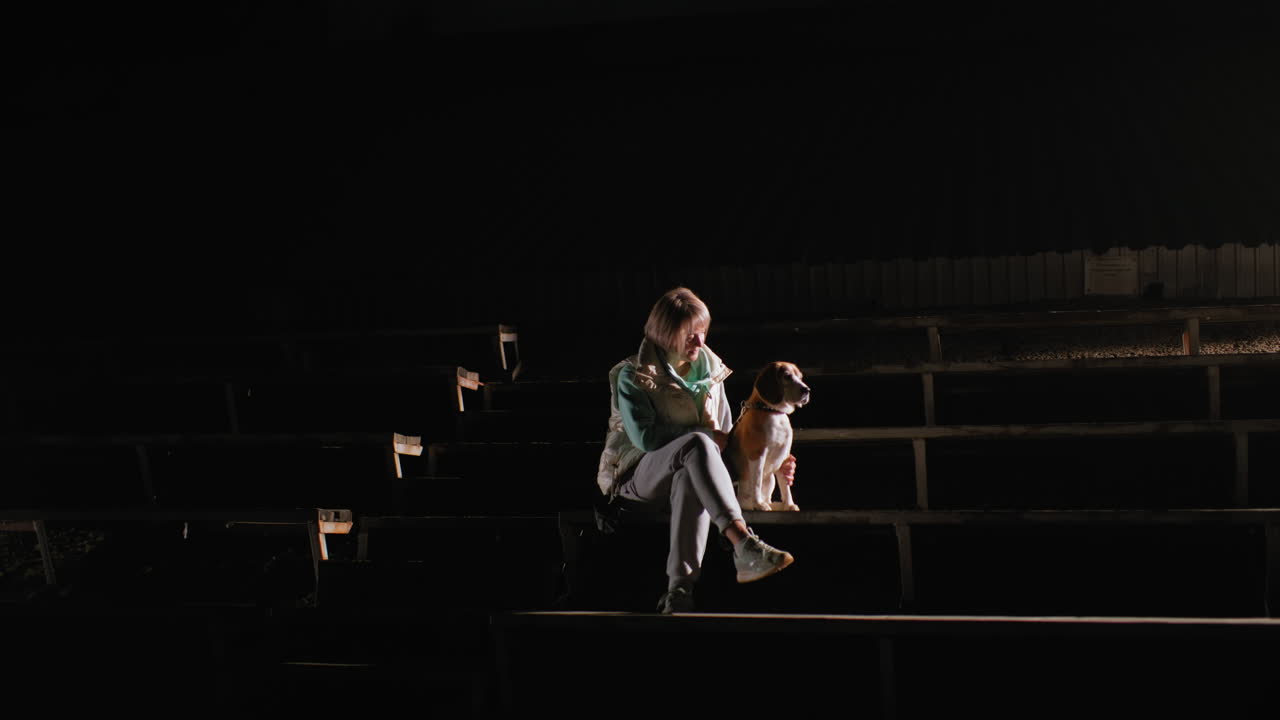 Sports woman seated on wooden stairs at night gently holding german shepherd puppy by her side, illuminated by spotlight in dark empty outdoor stadium creating peaceful and intimate atmosphere