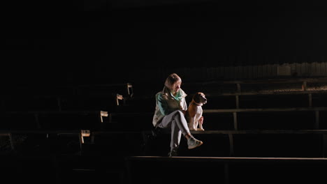 sports woman seated on wooden stairs at night gently holding german shepherd puppy by her side, illuminated by spotlight in dark empty outdoor stadium creating peaceful and intimate atmosphere