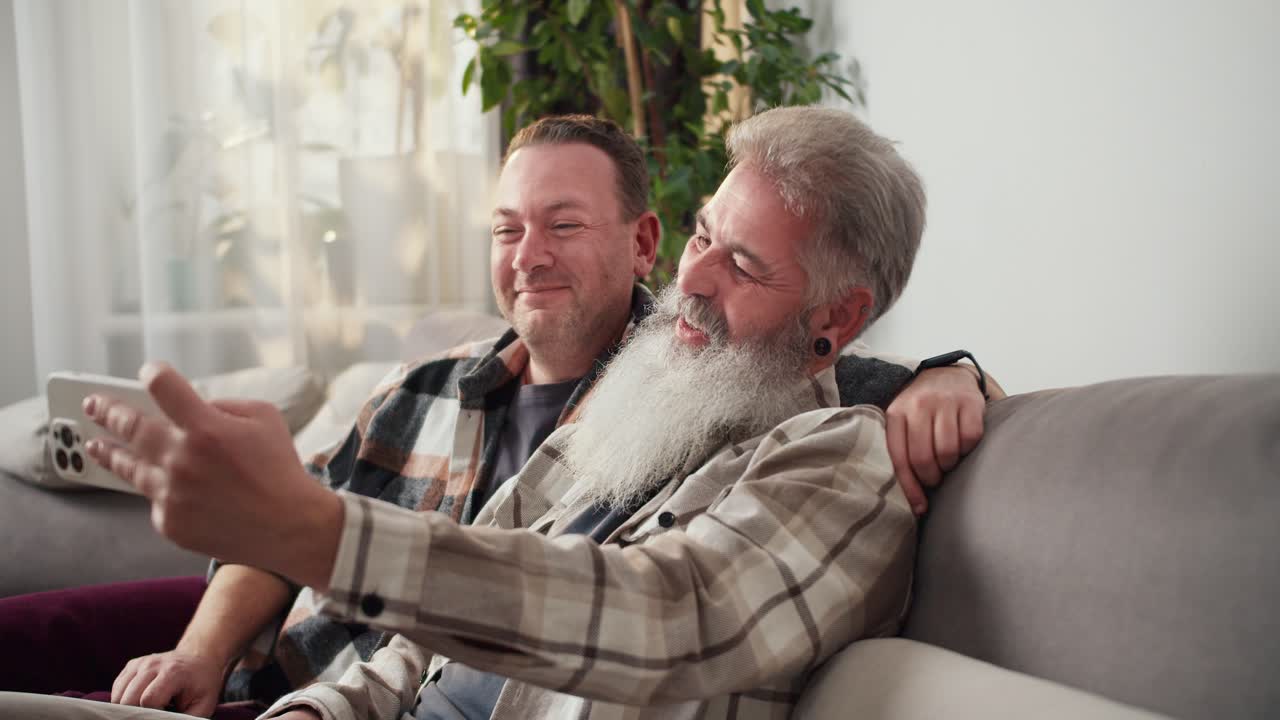 Happy middle-aged brunette man in a checkered shirt together with his elderly boyfriend with gray hair and beard takes a selfie using a White phone and communicates via video conference in a modern apartment on the sofa