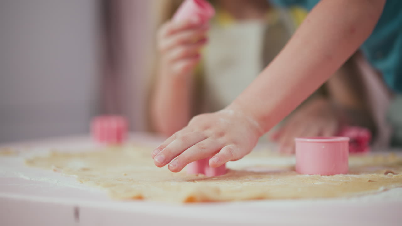 Close up of kid hand pressing pink cutter into flattened dough to create shapes, another kid blurred in background holding pink cutter, baking activity with flour spread over table