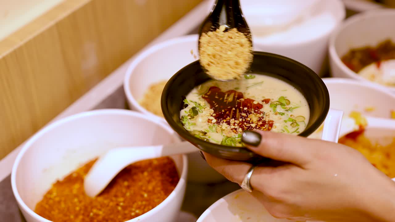 Hand sprinkles sesame and chili onto dipping sauce at self-service hot pot condiment station