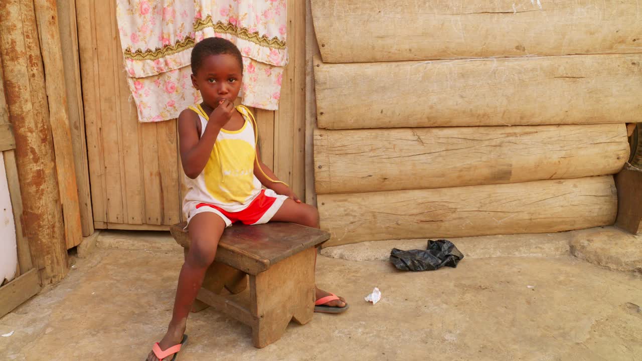 little cute black African girl sitting on a wooden chair while eating candy in her small remote rural village of Africa