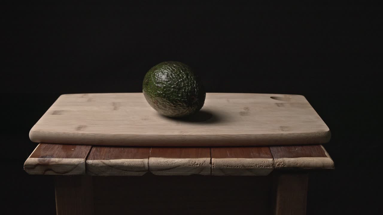 Un avocado maturo giace tranquillamente su un tavolo di legno invecchiato, circondato dall'oscurità. Un'illuminazione sottile e direzionale rivela la sua ricca consistenza e i suoi toni terrosi, creando un'immagine semplice ma potente.