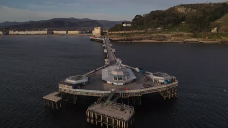 Vuelo-Aéreo-De-Drones-Alrededor-Del-Muelle-En-Llandudno-Gales-Mostrando-Las-Diversiones-Y-Una-Vista-Del-Paseo-Marítimo-Y-Las-Montañas