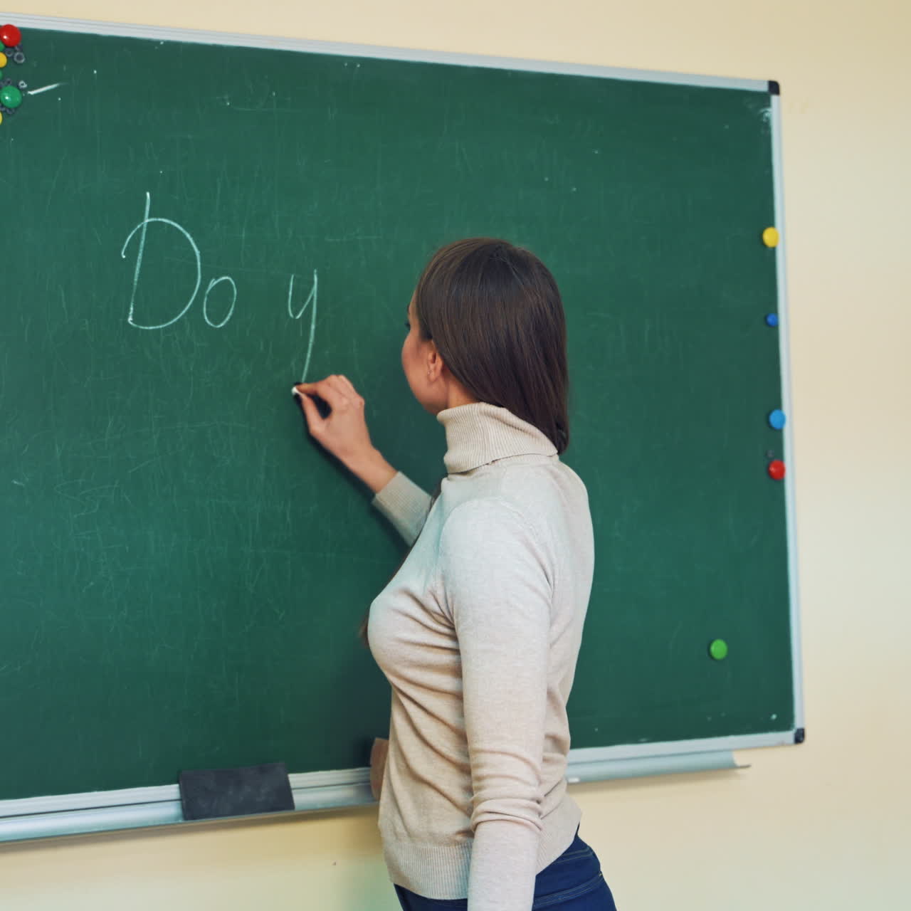 Smiling teacher stands near the blackboard in the classroom. Beautiful female teacher of English is writing a question on a blackboard with chalk. Educational concept.