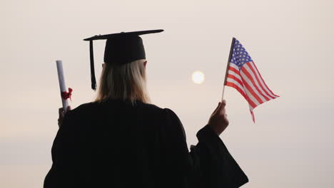 Ein-Student-In-Mantel-Und-Mütze-Hält-Ein-Diplom-Und-Die-Flagge-Der-Vereinigten-Staaten-Studiert-In-Amerika-United