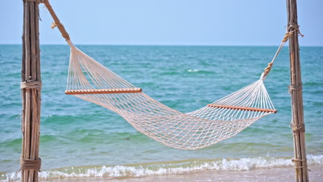 empty white hammock hangs from poles on tropical island beach with calm turquoise sea in background and tiny waves lapping at the sandy beach