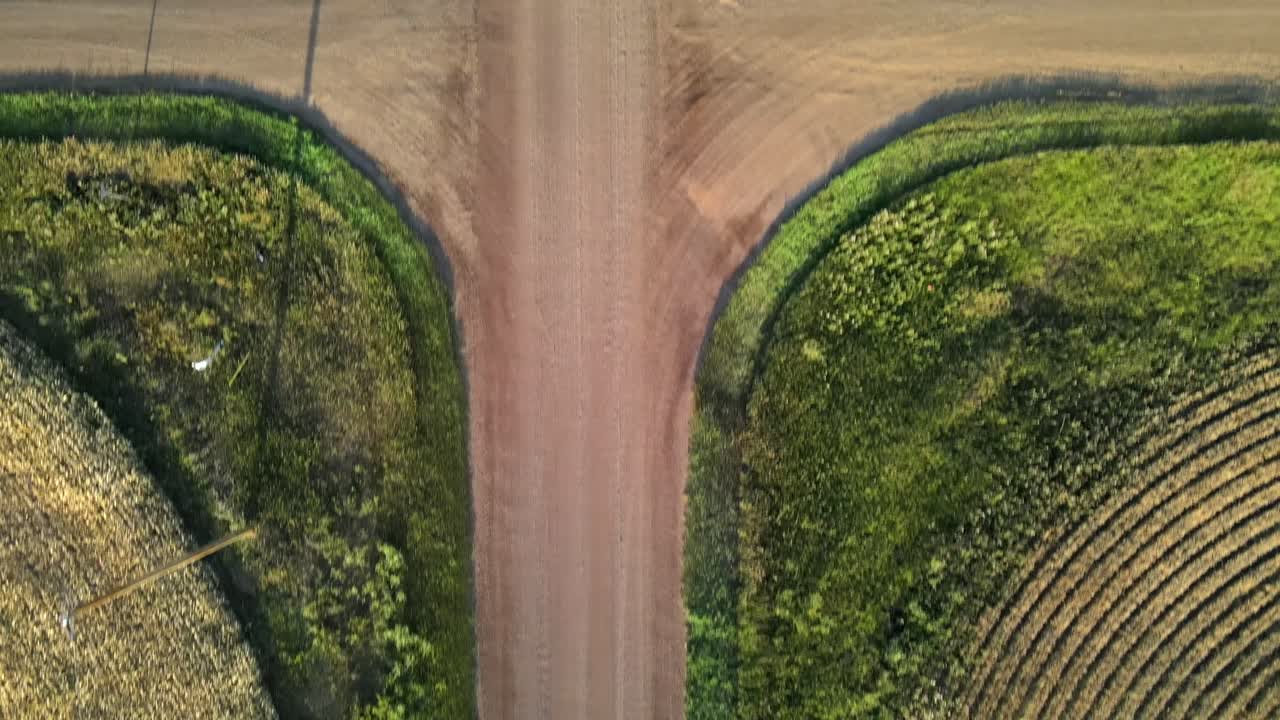 Toma aérea vertical en 4K mostrando un movimiento lento hacia adelante sobre una intersección de caminos de tierra roja entre campos de trigo y cebada en la Canadá rural.